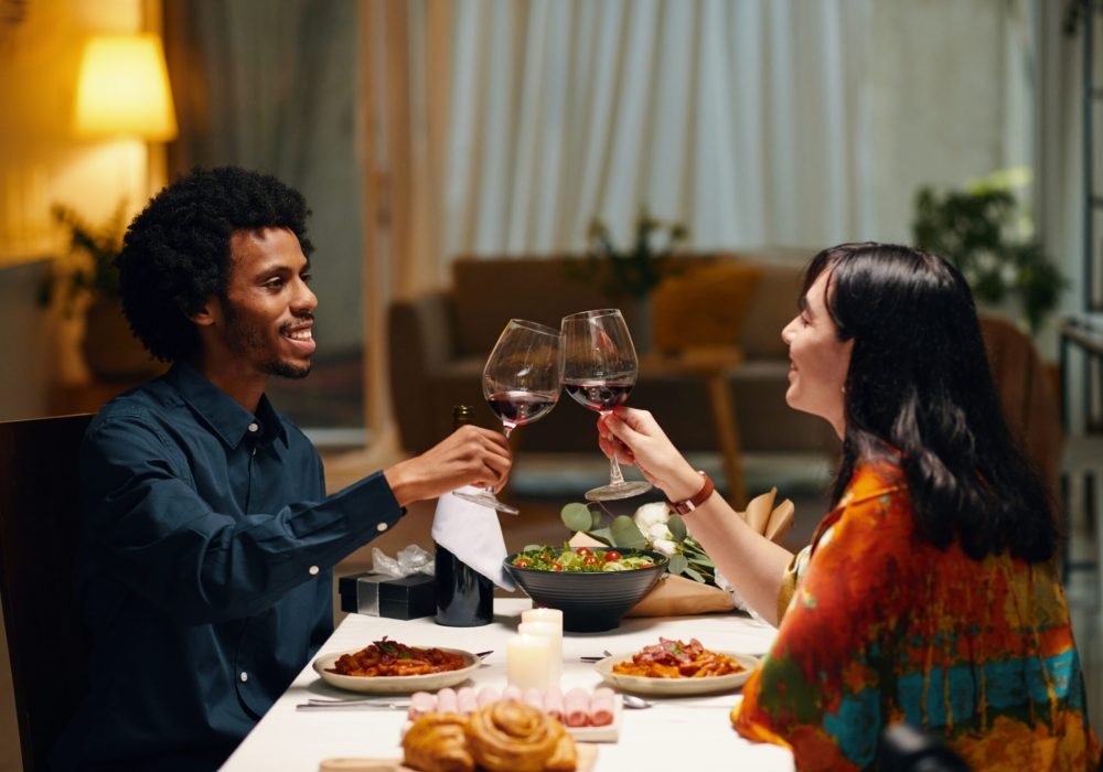 Side view of happy young intercultural valentines looking at one another and clinking with glasses of red wine over served festive table
