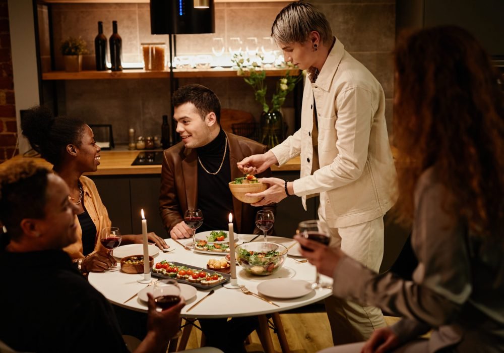 Young man putting vegetable salad on his plate while standing by dinner table served with homemade food among intercultural friends