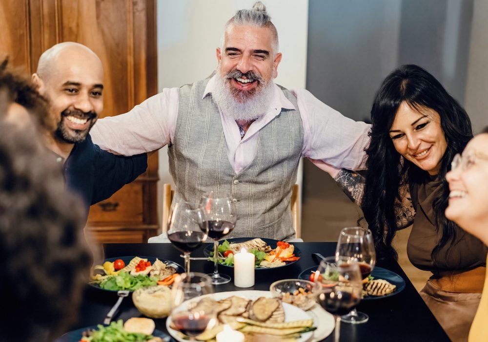 Adult cheerful friends hug happily, sitting at the table during a dinner. Selective focus on the bearded man in the center - high iso image.