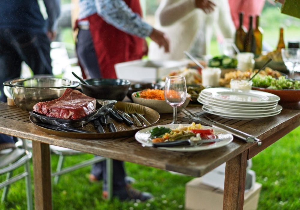 Close-up of a rustic wooden table laden with food for an outdoor barbecue party. Raw steak, salads, wine glasses, and plates are visible with people in the background enjoying the gathering.