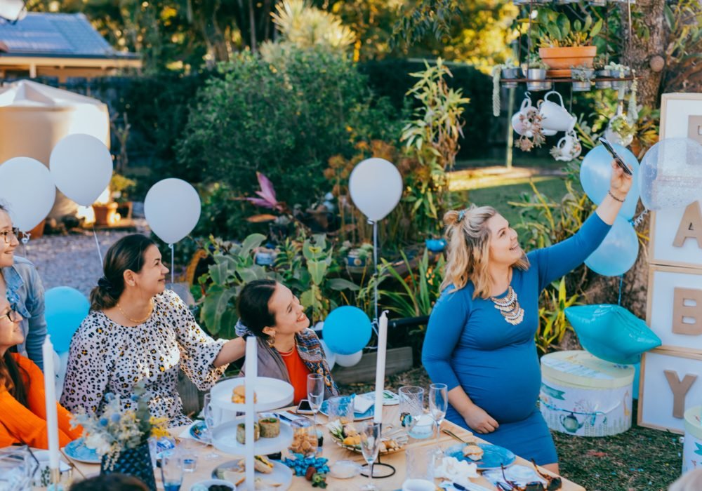Female friends taking selfie with pregnant woman at a baby shower. Mobile photography, party decorations in white and blue colors, baby boy
